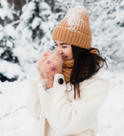 A beautiful happy laughing young girl in gloves and a winter hat-scarf, covered with snow flakes. Winter forest landscape backgroundの写真素材