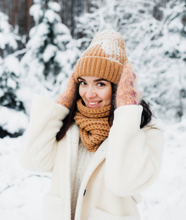 A beautiful happy laughing young girl in gloves and a winter hat-scarf, covered with snow flakes. Winter forest landscape backgroundの写真素材