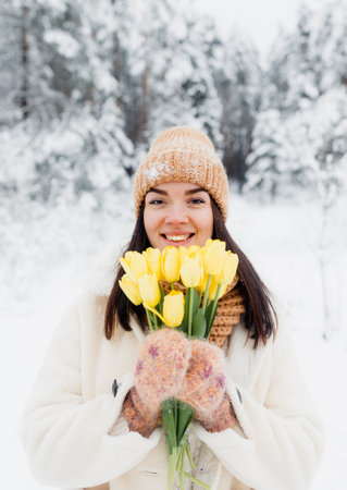 A beautiful happy young girl in gloves and a winter hat-scarf holds a bouquet of yellow tulips. Winter forest landscape backgroundの写真素材