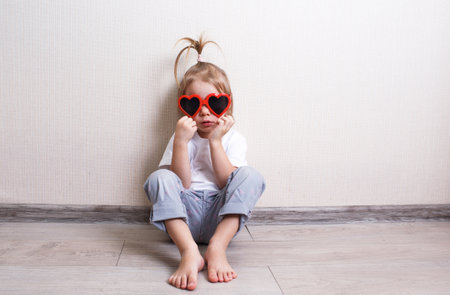 a little girl wearing sunglasses is sitting on the floor of a room against the wall.の写真素材