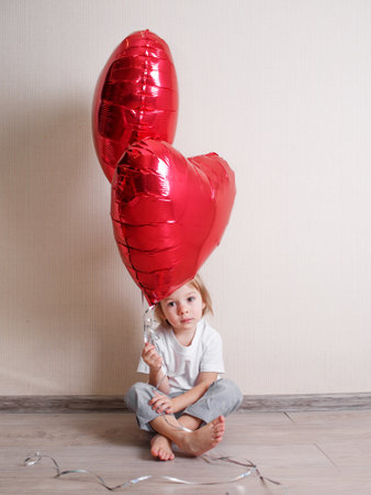 a little beautiful girl is sitting at home on the floor with red heart-shaped balls in her hands.の写真素材