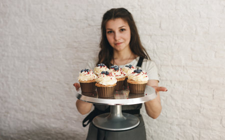 The cook girl holds a tray with beautiful cupcakes decorated with cream and berries.の写真素材