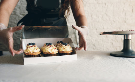 A girl cook in a gray apron packs cupcakes with cream in a gift box to send the order to the customer.の写真素材