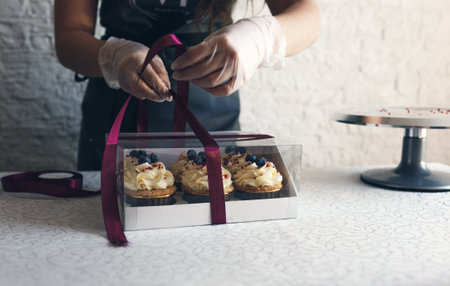 A girl cook in a gray apron packs cupcakes with cream in a gift box to send the order to the customer.の写真素材