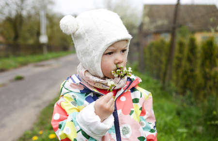 a little girl in a bright jacket stands outside in the spring and holds a branch with spring white flowers in her hands.の写真素材