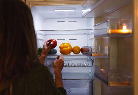 a womans hand takes a tomato from the refrigerator shelf. The refrigerator is filled with vegetables and fruits. healthy food. healthy lifestyle conceptの写真素材