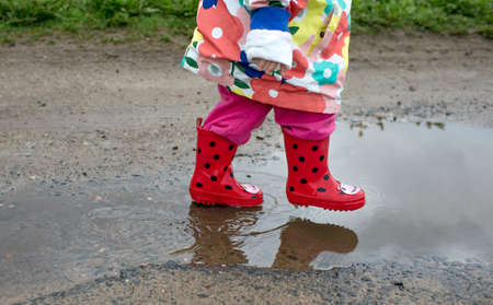 A little girl in a bright jacket and red boots jumps merrily through puddles. Spring mood.の写真素材