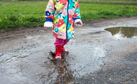 A little girl in a bright jacket and red boots jumps merrily through puddles. Spring mood.の写真素材