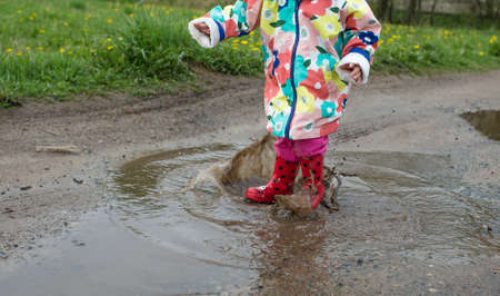 A little girl in a bright jacket and red boots jumps merrily through puddles. Spring mood.の写真素材