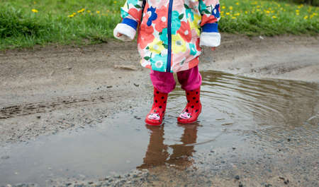 A little girl in a bright jacket and red boots jumps merrily through puddles. Spring mood.の写真素材