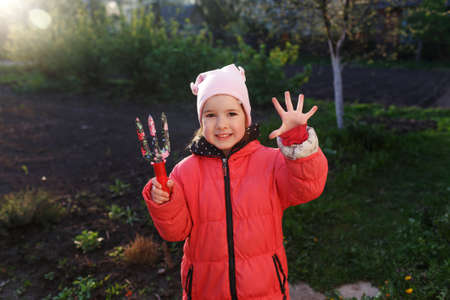 a little girl in a red jacket is standing in the garden with gardening supplies. Children help their parents in their work.の写真素材