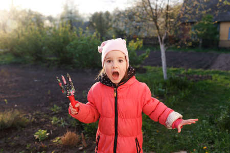 a little girl in a red jacket is standing in the garden with gardening supplies. Children help their parents in their work.の写真素材