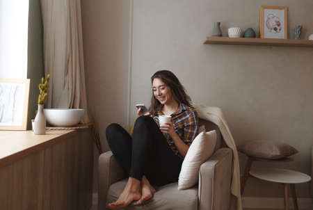 A young woman with a mobile phone is sitting in a comfortable chair by the window of the house and drinking tea. Home interior design. Freelancerの写真素材