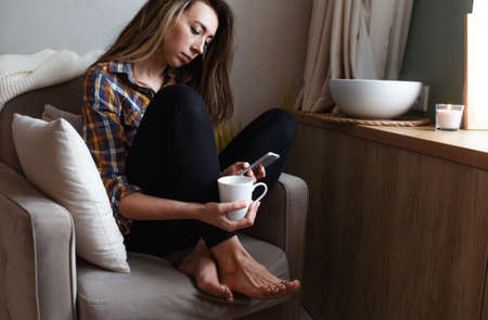 A young woman with a mobile phone is sitting in a comfortable chair by the window of the house and drinking tea. Home interior design. Freelancerの写真素材