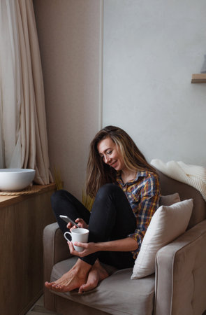 A young woman with a mobile phone is sitting in a comfortable chair by the window of the house and drinking tea. Home interior design. Freelancerの写真素材