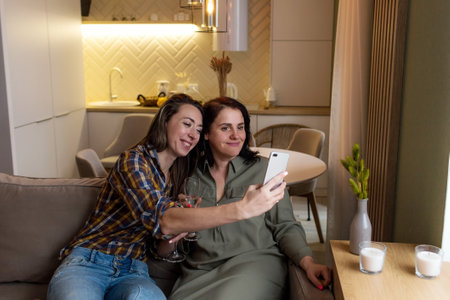 Two girls are sitting on the couch, raising their glasses and taking selfies on their phone.の写真素材
