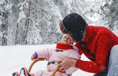 A young father rides a wooden sled with his adorable daughter in a snow-covered forest.Childrens games in the snowy forest. Family winter vacation with a childの写真素材