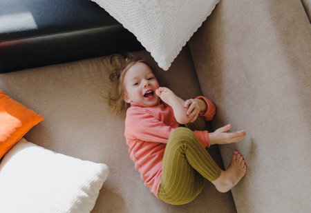 a little girl in a pink sweatshirt is lying on the sofa among the pillows with her legs up. Home games.の写真素材