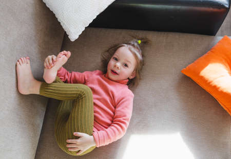 a little girl in a pink sweatshirt is lying on the sofa among the pillows with her legs up. Home games.の写真素材
