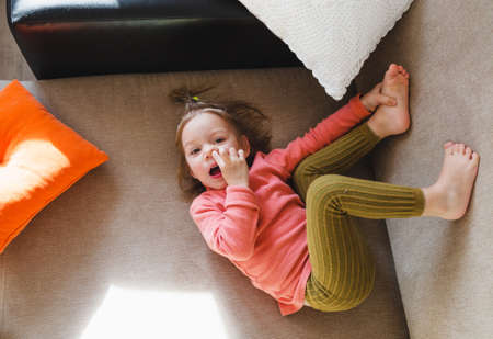 a little girl in a pink sweatshirt is lying on the sofa among the pillows with her legs up and picking her nose. Home games.の写真素材