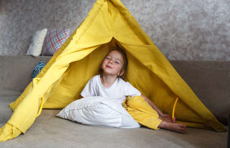 A beautiful girl in yellow pants and a white T-shirt smiles and plays inside the teepee at home on the couchの写真素材