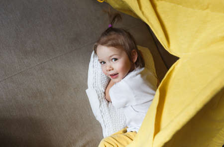 A beautiful girl in yellow pants and a white T-shirt smiles and plays inside the teepee at home on the couch. Top view.の写真素材
