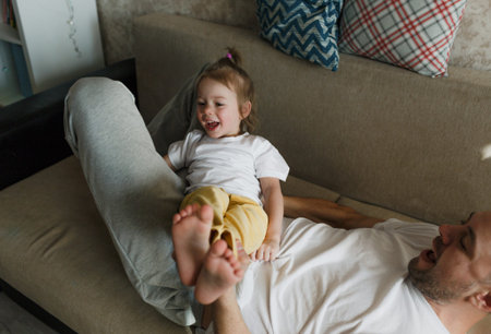 a little girl in a white T-shirt is having fun playing with her dad on the couch, having fun enjoying each other.の写真素材