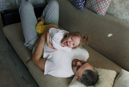 a little girl in a white T-shirt is having fun playing with her dad on the couch, having fun...