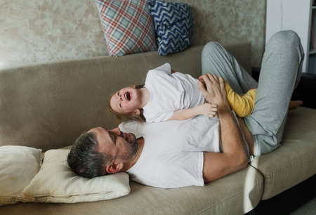 a little girl in a white T-shirt is having fun playing with her dad on the couch, having fun enjoying each other.の写真素材