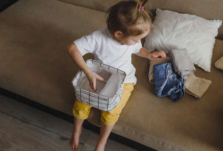 a little girl is sitting on the sofa and neatly folding clothes in a storage box. Moms assistantの写真素材
