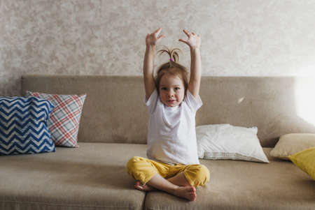 a little girl is sitting on the couch in yellow pants and a white T-shirt and doing morning exercises.の写真素材
