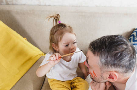 A little girl on the couch paints her fathers face during family games together.の写真素材