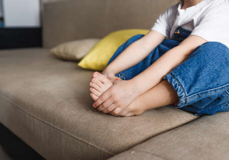 a little girl in a blue denim jumpsuit is sitting on the couch with her arms wrapped around her legs. Close-up.の写真素材