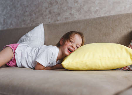 a girl in a white T-shirt is lying at home on the couch and laughing with her head on a yellow pillow.の写真素材