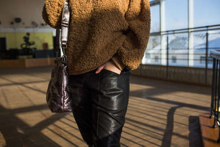 a girl in a brown sweater, black trousers with a large womens bag holds her hand in her pocket against the background of large windows. Close-upの写真素材