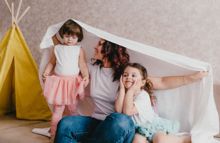 a happy young mother and two small daughters play together on the sofa covered with a sheet. happy family relationships.の写真素材