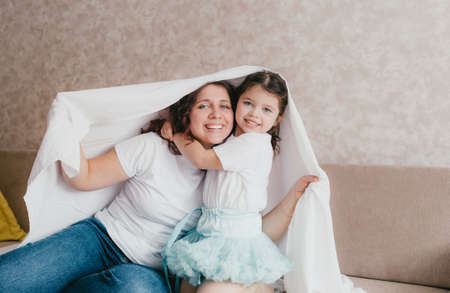 a happy little girl in a light turtleneck hugs her mother under a white sheet. a loving and caring motherの写真素材