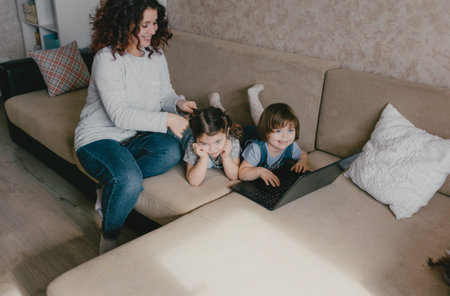 two little girls play on a laptop while lying on the couch mom does her daughters hair.の写真素材