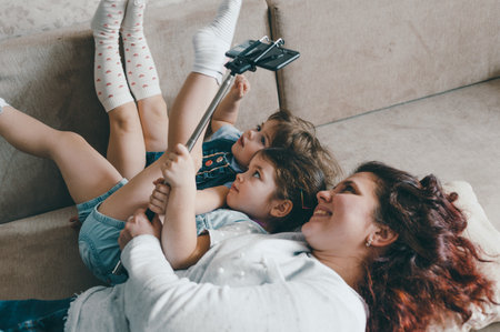 happy mom with two young daughters lies on the couch and takes pictures of herself on the phoneの写真素材