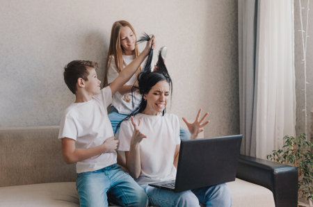 An irritated mother sits on the couch and works at home on a laptop, active children play with their mothers hair, do her hair. Tired, stressed mom cant concentrateの写真素材