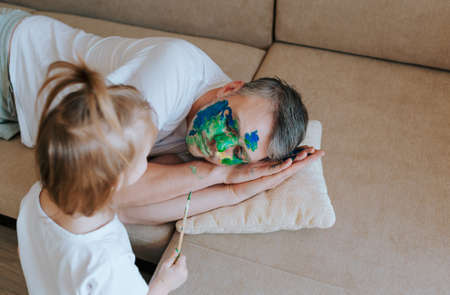 A little girl paints her fathers face with a brush and watercolors while he sleeps on the couchの写真素材