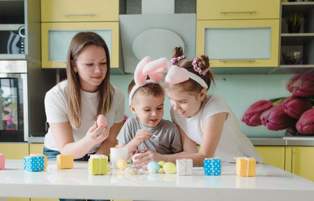 Happy family: mother daughter and son with bunny ears are preparing for the holiday, coloring eggs in the cozy kitchen of the house. preparations for the Easter holidayの写真素材