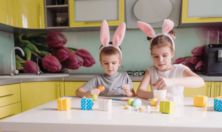 a young brother and sister with rabbit ears on their head paints Easter eggs at home in the kitchen....