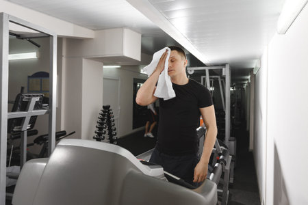 An exhausted man wipes his sweaty face with a hand towel after an intense workout at the gym on the...