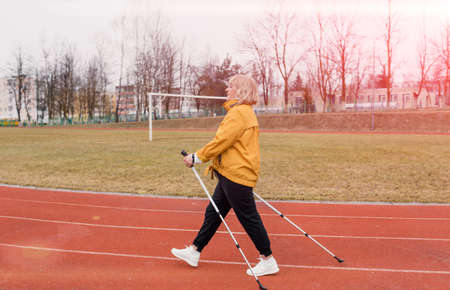 An elderly woman in a yellow sports jacket practices Nordic walking outdoors on the stadium's rubber treadmill. A sunny sunset. Older women walk with a Nordic gait, using trekking poles and Nordic poles. Pensioners healthy lifestyleの写真素材