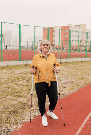 An elderly woman in a yellow jacket stands with Nordic sticks in her hands at the stadium on a red treadmill. healthy lifestyleの写真素材
