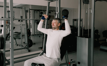 Portrait of a young man doing physical exercises, training with dumbbells sitting on a bench in the...