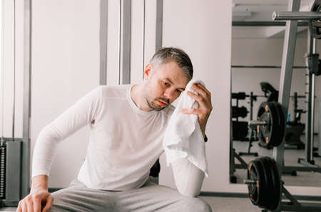 a young man tired after a hard workout sits and wipes the sweat from his face with a towel at the sports club. rest recovery.の写真素材