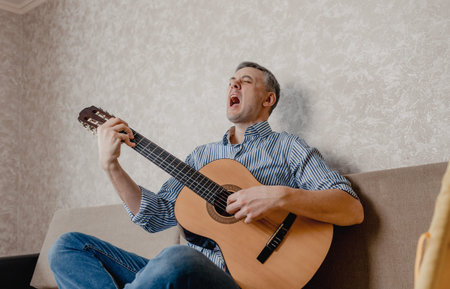 A young man plays the guitar sitting on a sofa in a bright living room. A musical instrument for the...