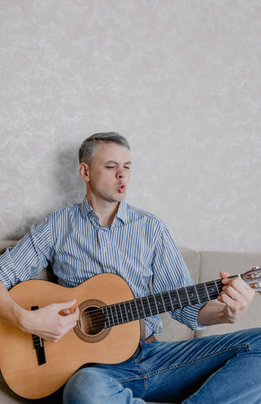 A young man plays the guitar sitting on a sofa in a bright living room. A musical instrument for the...